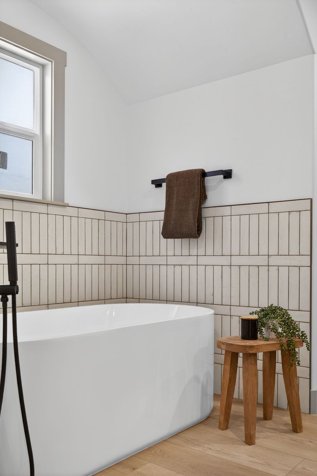 Bright bathroom with freestanding tub, tiled walls, brown towel, and wooden stool with plant.