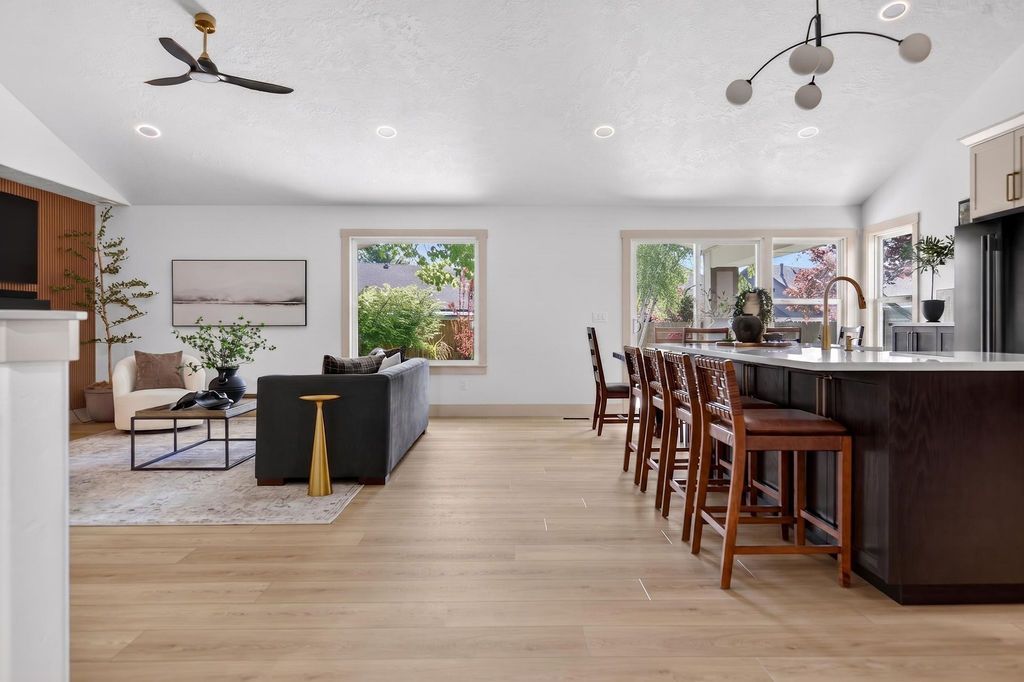 Modern open-concept living room and kitchen with wood floors, gray sofa, bar stools, and large windows