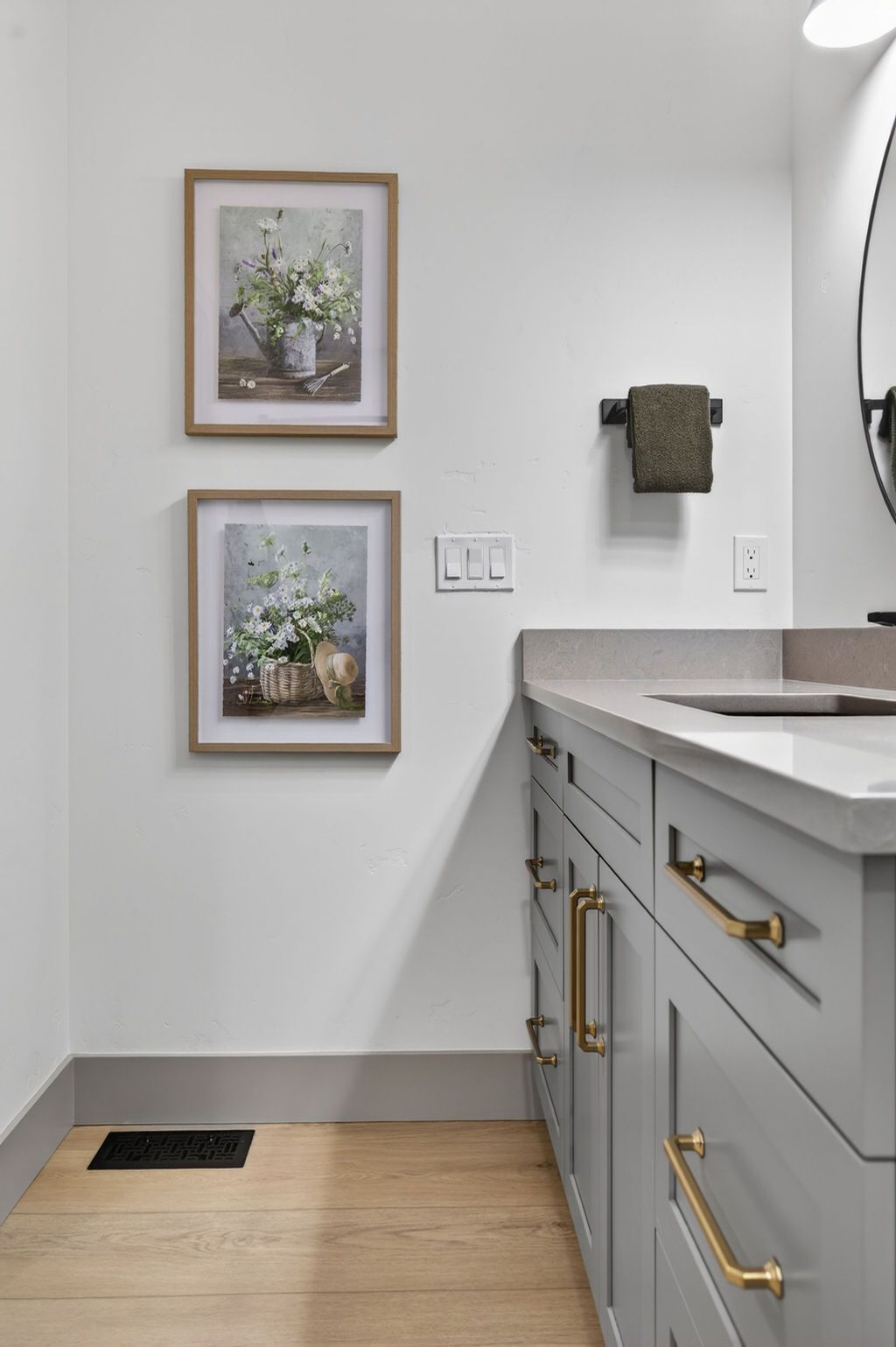 Bathroom vanity with gray cabinets, brass pulls, framed floral prints, and a towel ring on a white wall