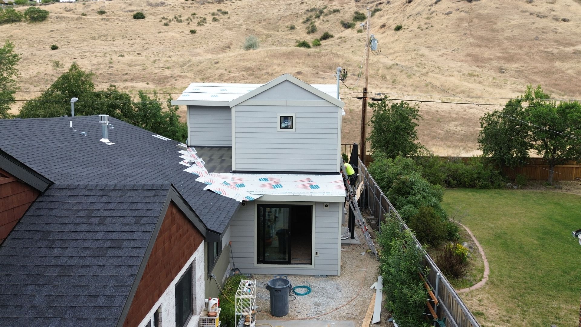 Rear view of a small white house with dark roof, a back deck, and hillside yard with a fence