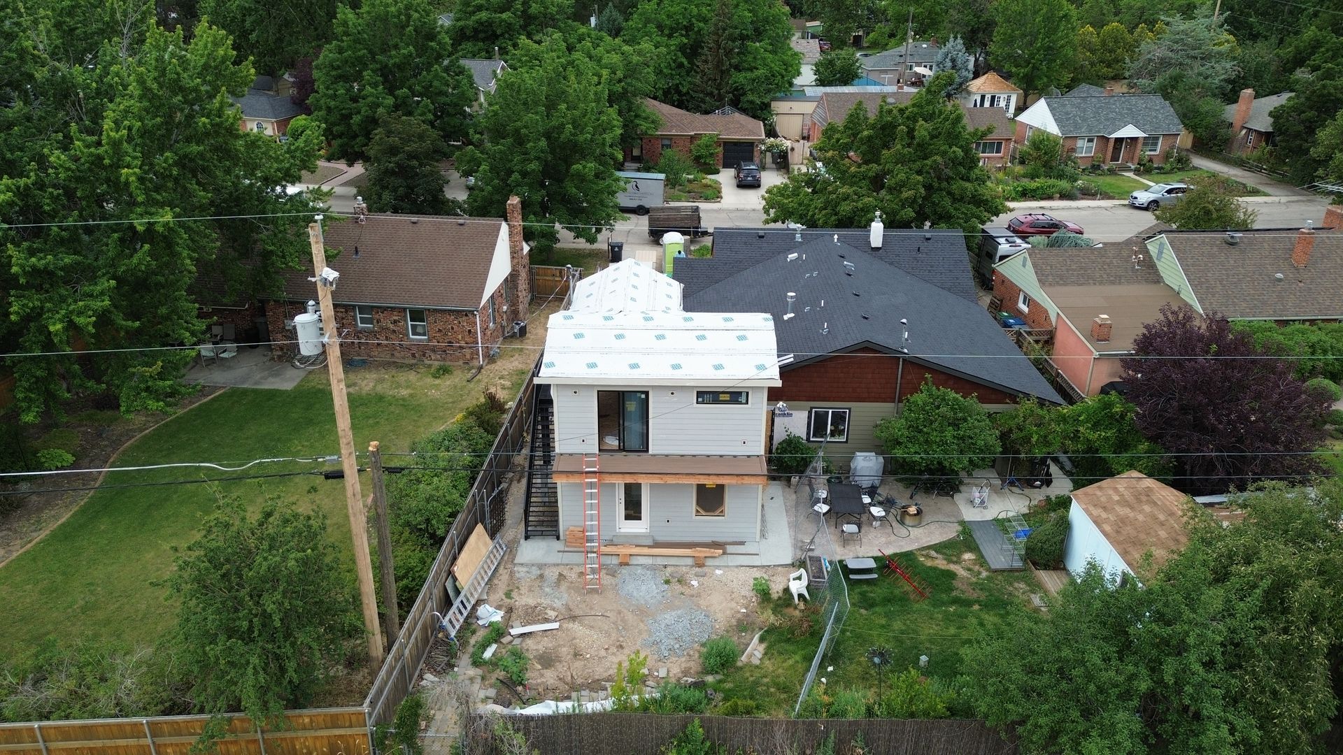 Aerial view of a backyard with a white house, patio, fence, and neighboring homes surrounded by trees.
