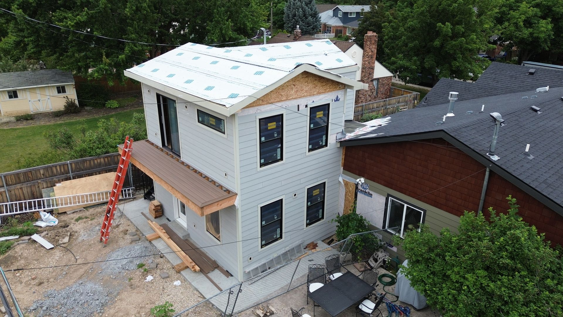 New white two-story house under construction beside a red brick home, with ladders and roof work visible