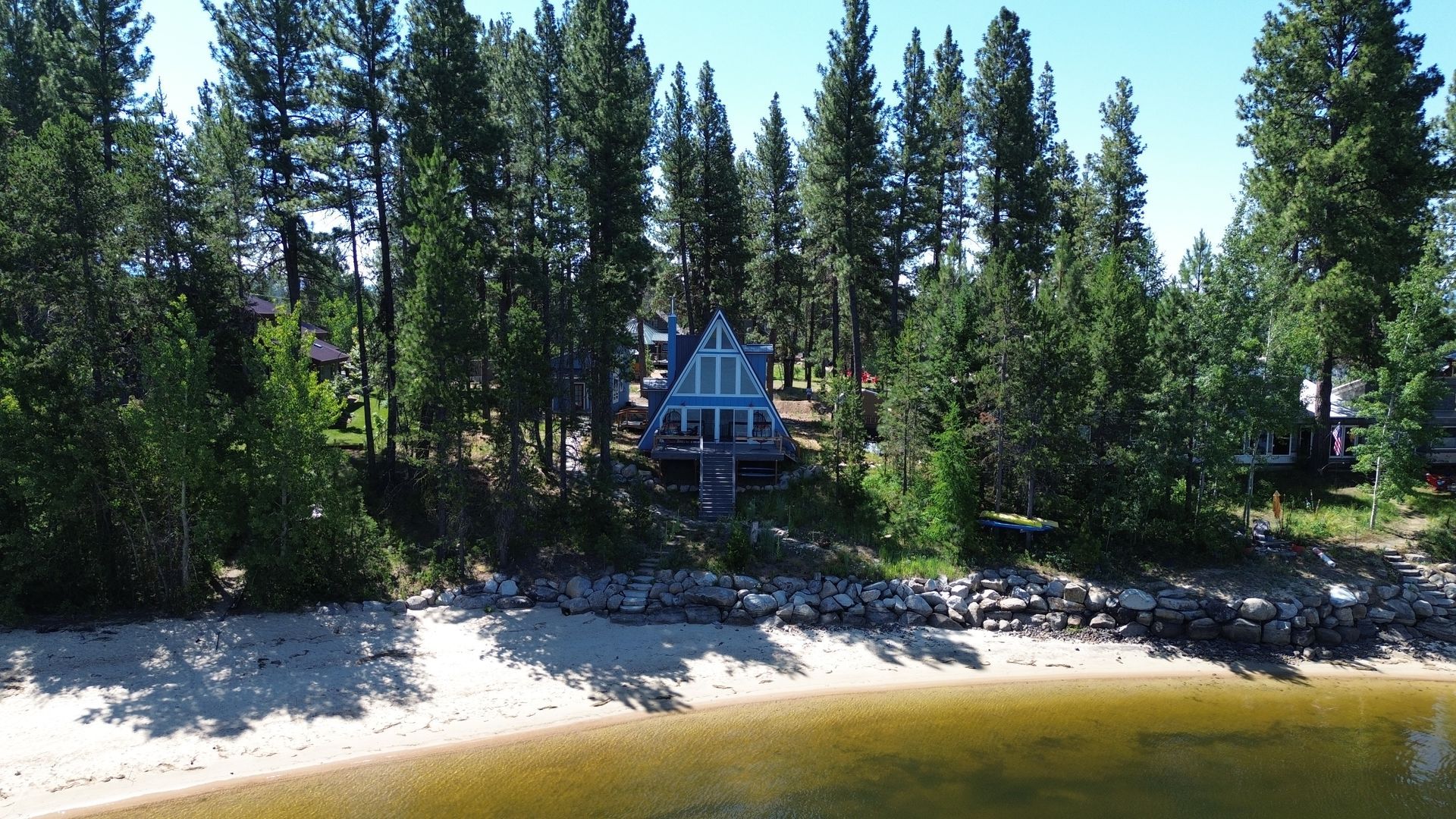 Lakeside A-frame cabin among tall pine trees, with a sandy shore and calm water in front.