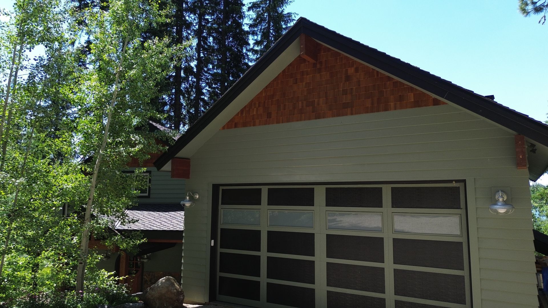 Small house with white siding, red gable, and garage doors under tall trees in daylight