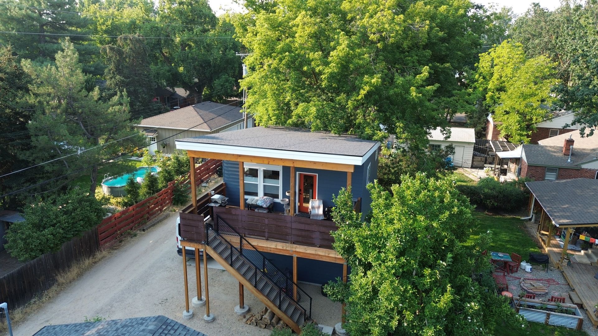 Aerial view of a small house with a wooden deck, surrounded by trees and a fenced yard.