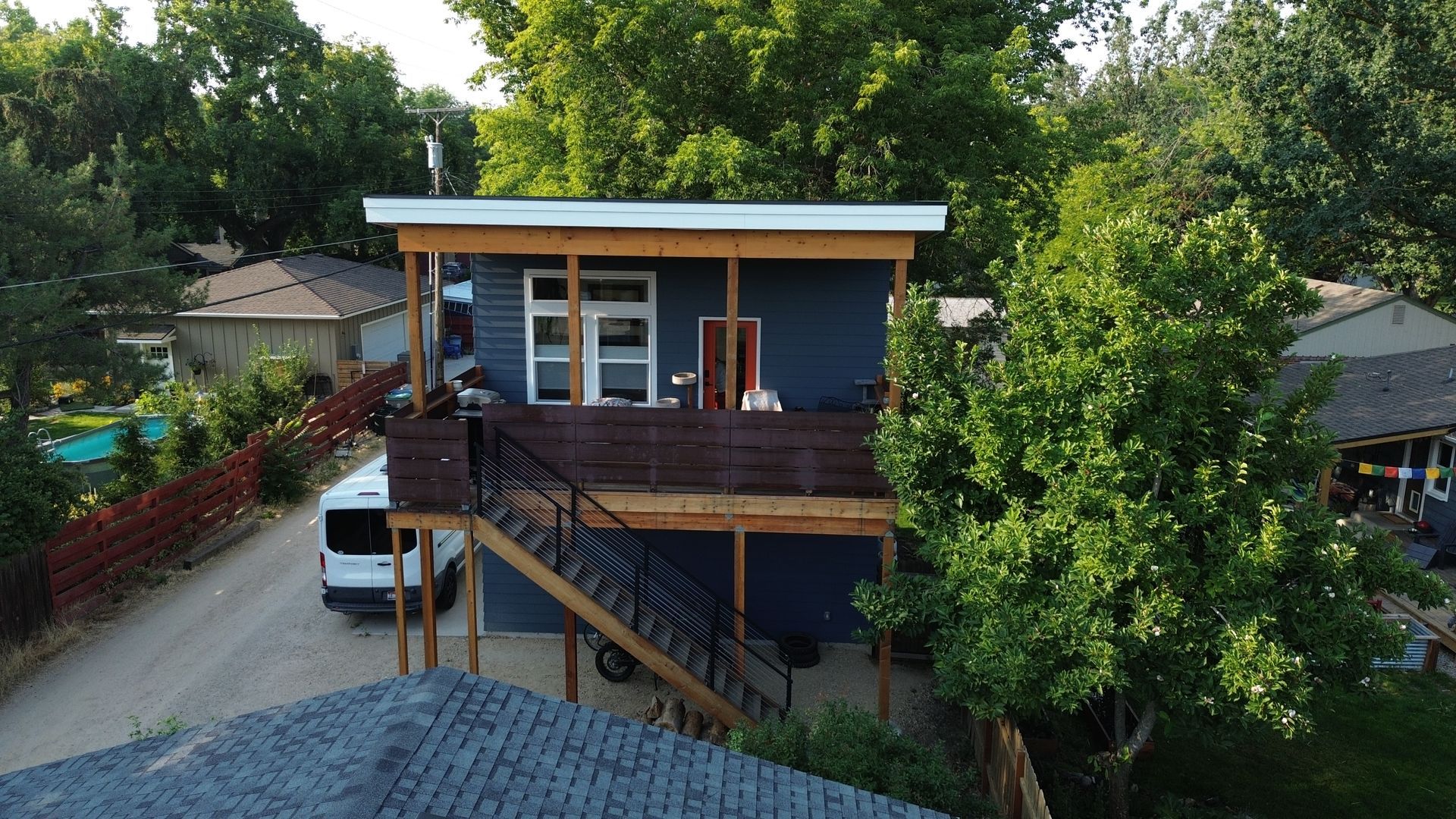 Treehouse-style house on stilts with a rooftop deck, surrounded by tall green trees and neighboring homes.