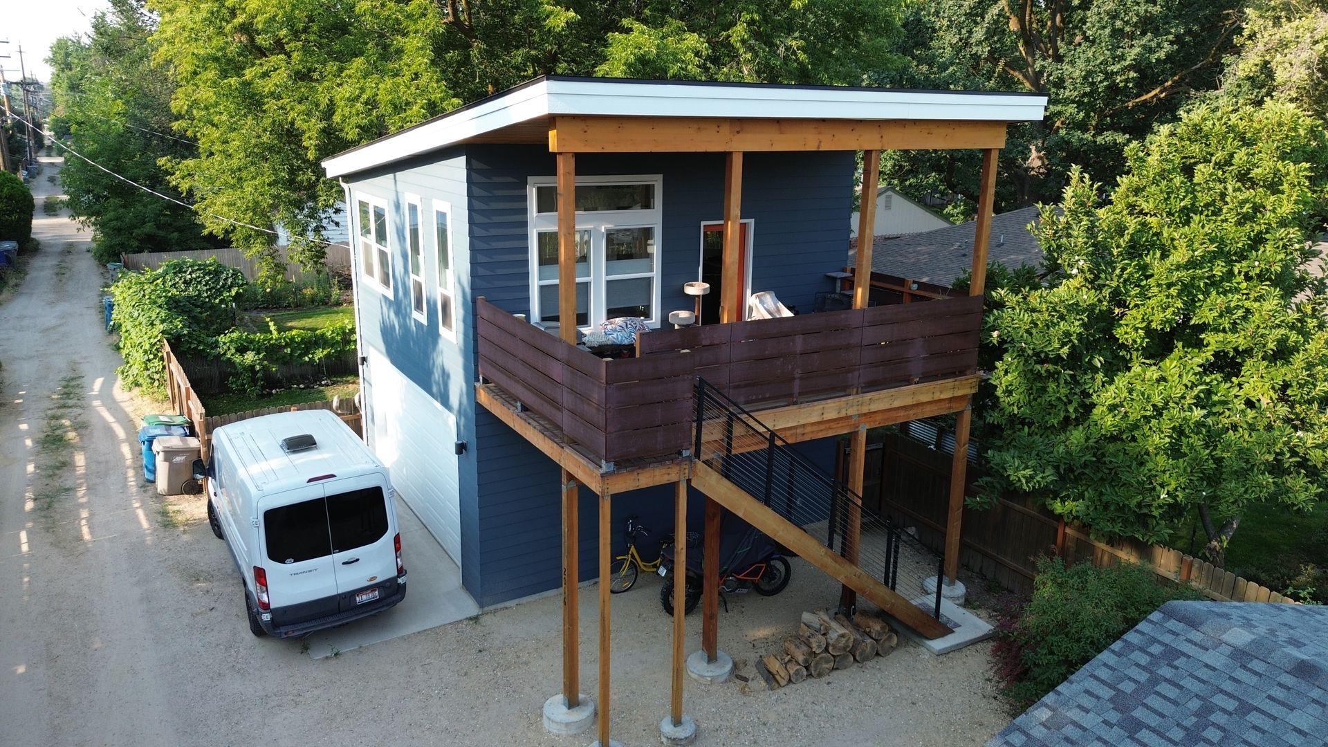 Modern two-story house with wooden balcony, stairs, and a white van parked beside it