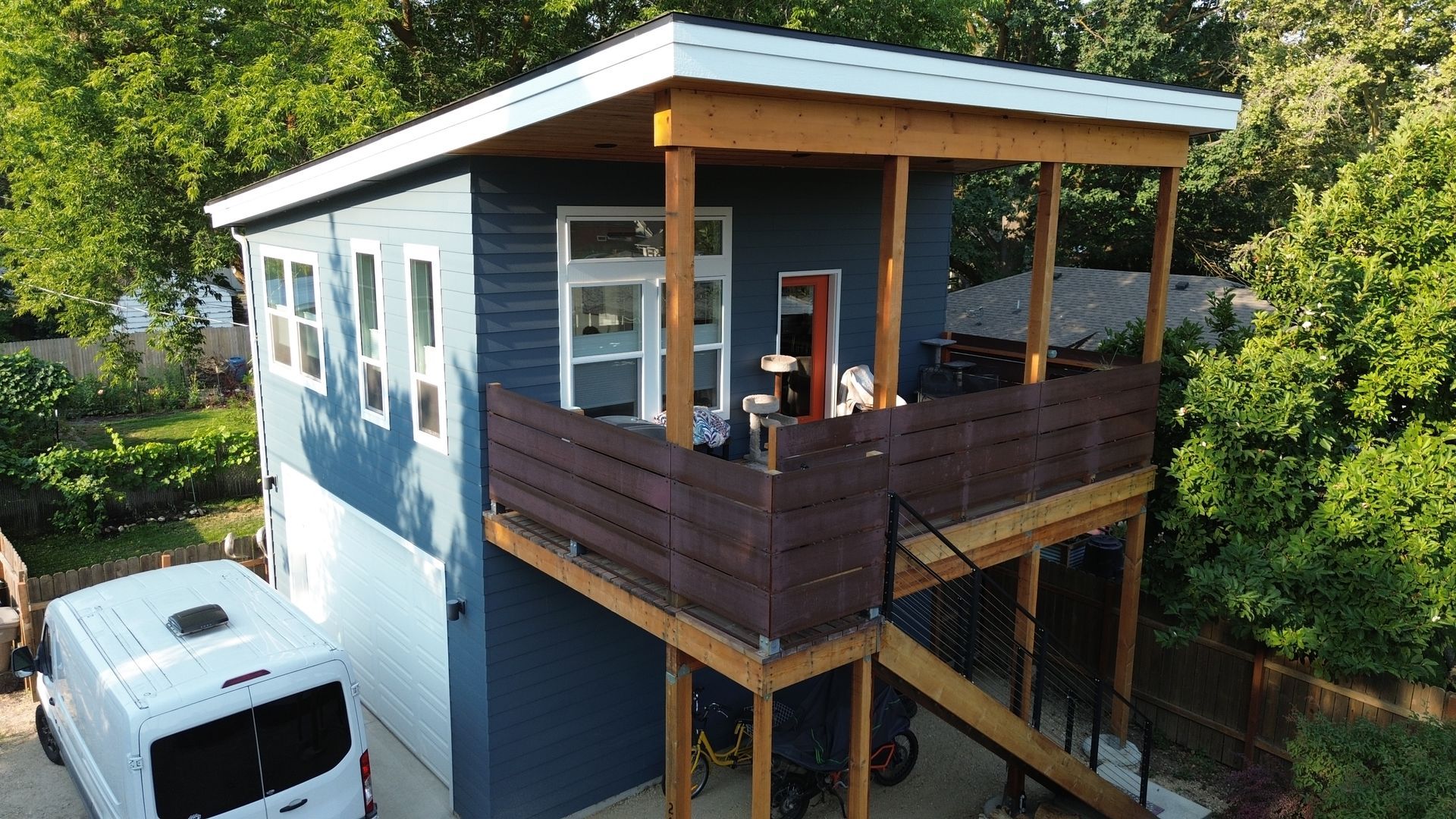 Modern two-story house with a rooftop deck and white camper parked beside it, surrounded by trees.