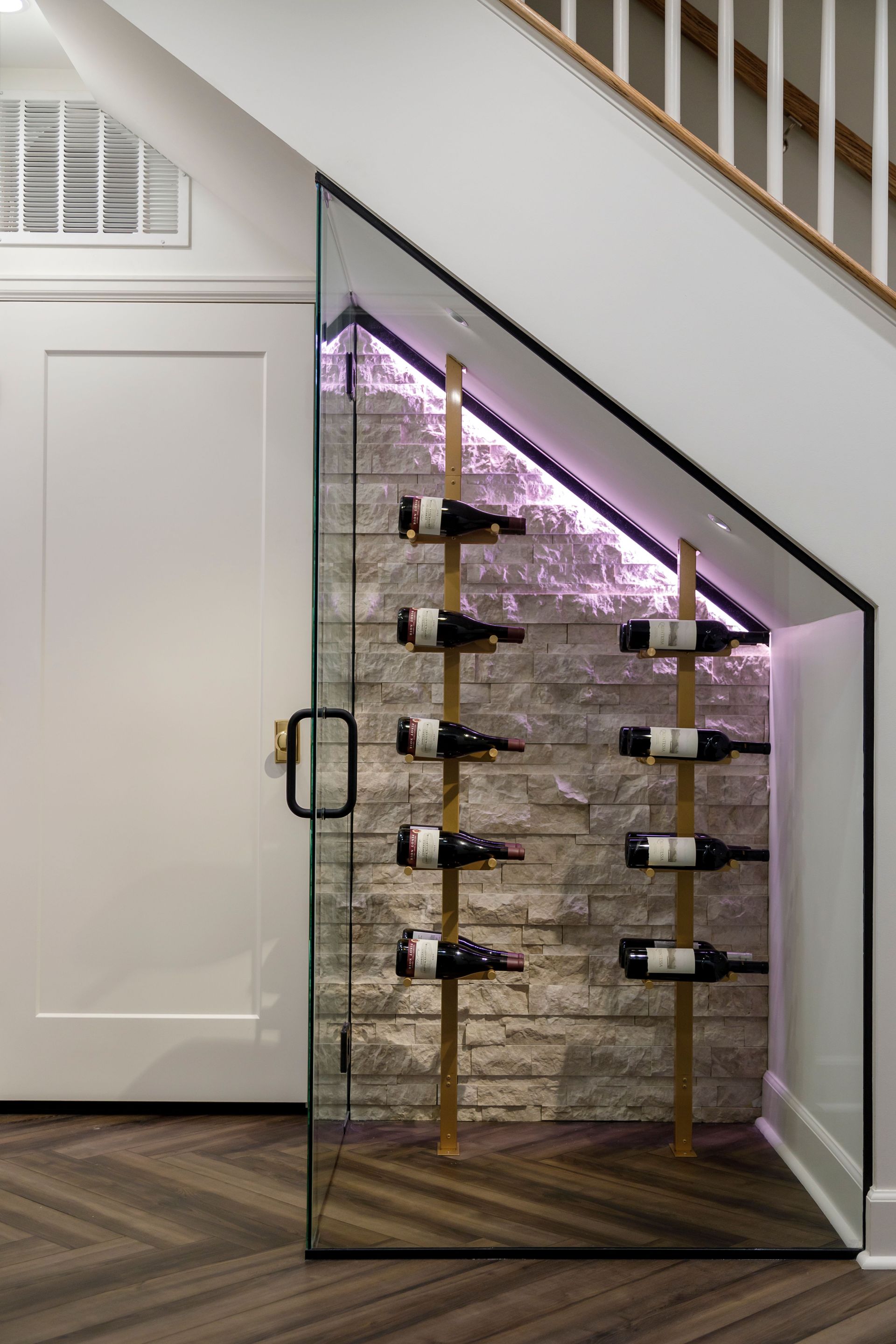 Glass-front wine storage under a staircase with bottles on metal racks and purple lighting