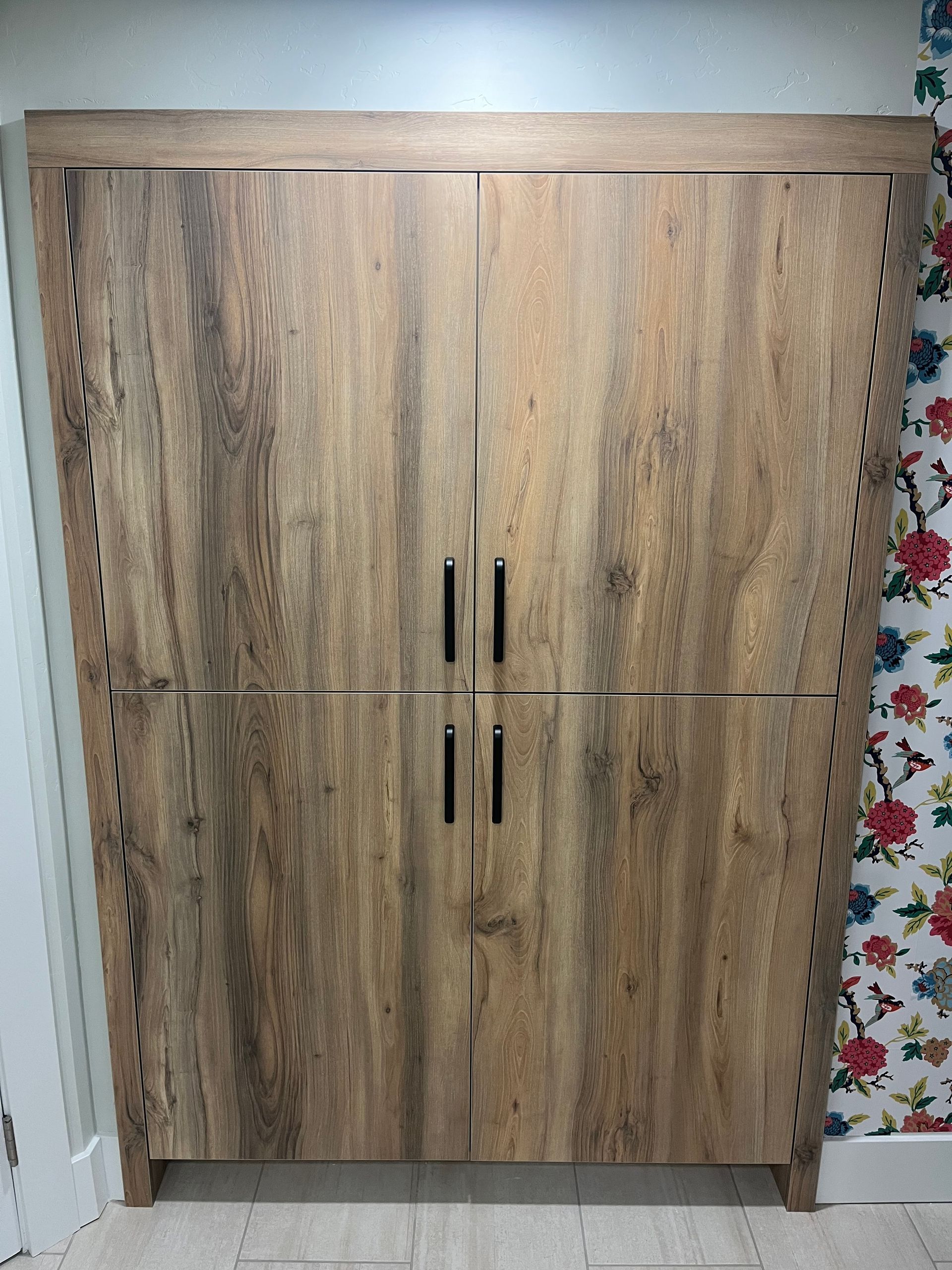 Wood-grain wardrobe cabinet with four doors against a wall, beside floral wallpaper.