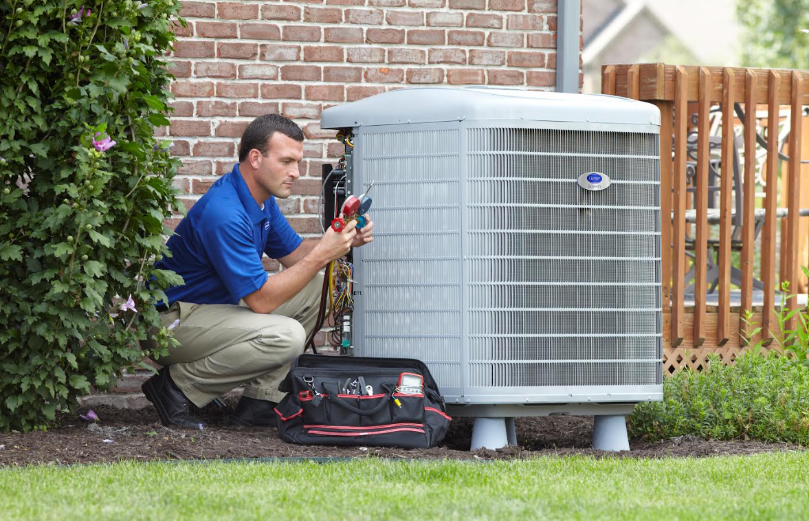 Technician kneeling beside an outdoor air conditioner, using tools in a backyard.