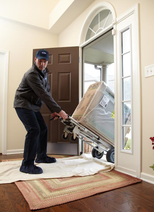 Mover carrying a wrapped appliance through a front doorway onto a rug