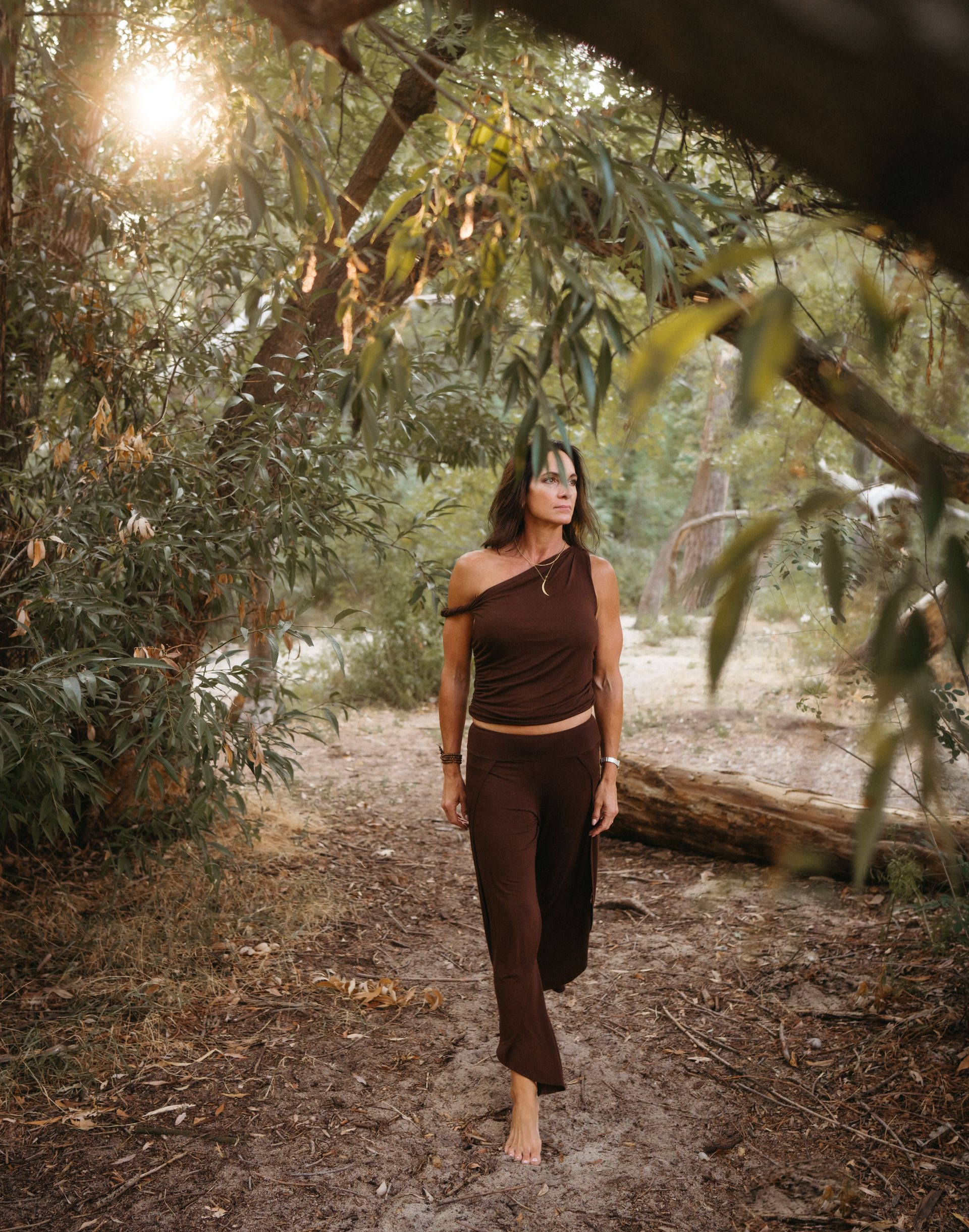 Nicole  in flowing brown yoga outfit walking barefoot through the trees with the sun peaking through