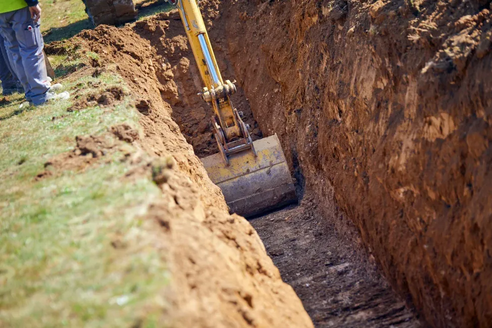 A yellow excavator bucket digs a deep, narrow trench in a grassy, dirt-filled field.