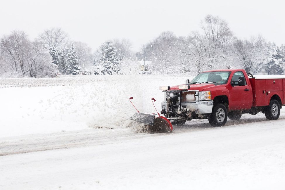 A red pickup truck with a snowplow blade attached moves through a snowy field, spraying snow as it clears the ground.
