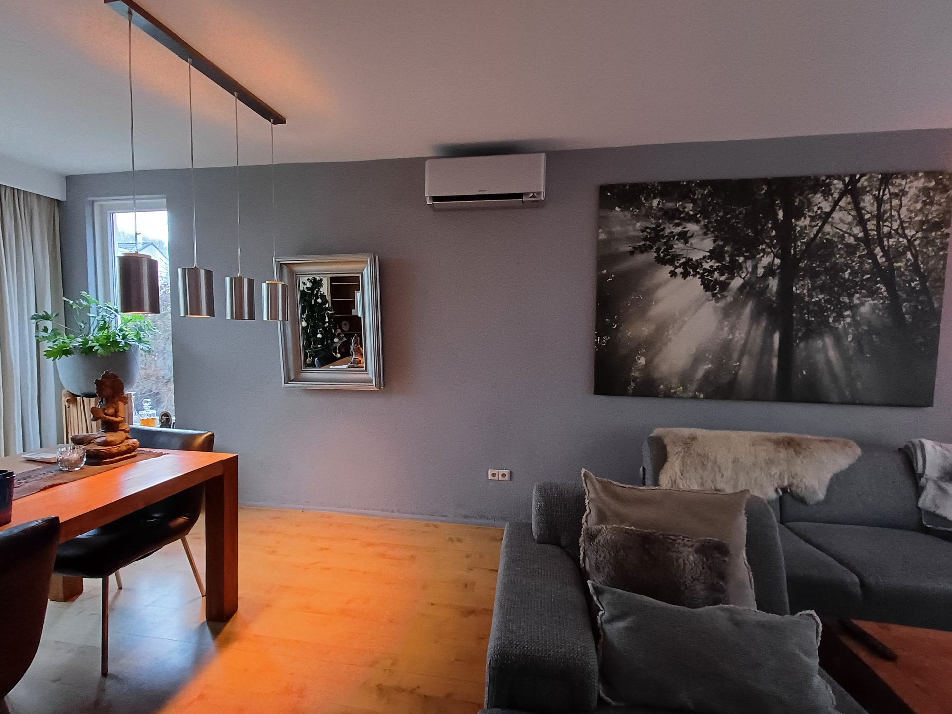 Moderne woonkamer met grijze muren, houten vloer, een Daikin Stylish airco aan de muur, een grote zwart-wit wandfoto van een bos, een grijze hoekbank en een houten eettafel met hanglampen en decoraties.