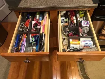 Two open wooden kitchen drawers with plastic organizers filled with various household items and office supplies.