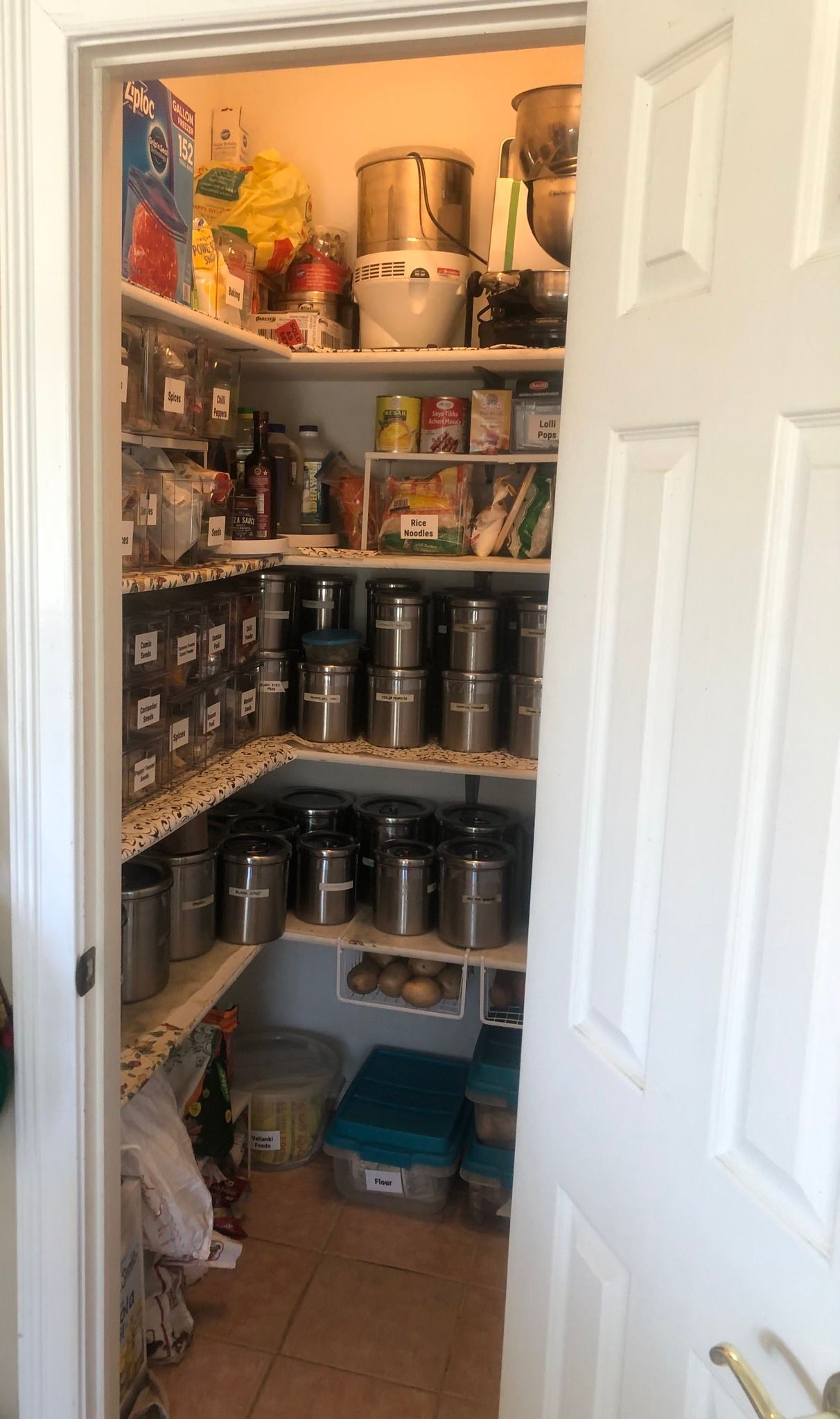 Pantry interior filled with shelves, storage containers, and various food items, accessible from an open white door.