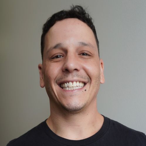 Man smiling, wearing a dark shirt, in front of a wooden wall.