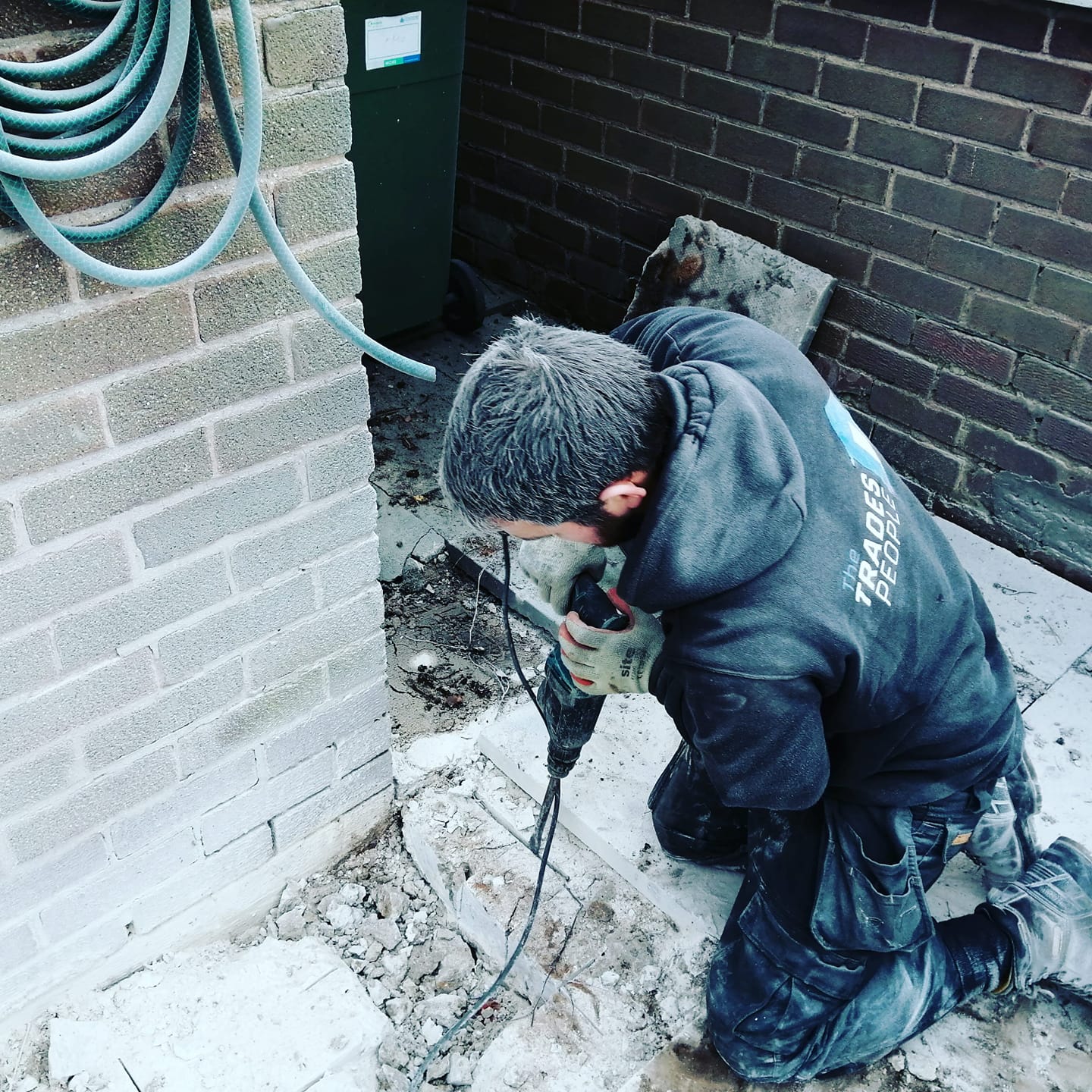 A man is kneeling down using a drill in front of a brick wall.