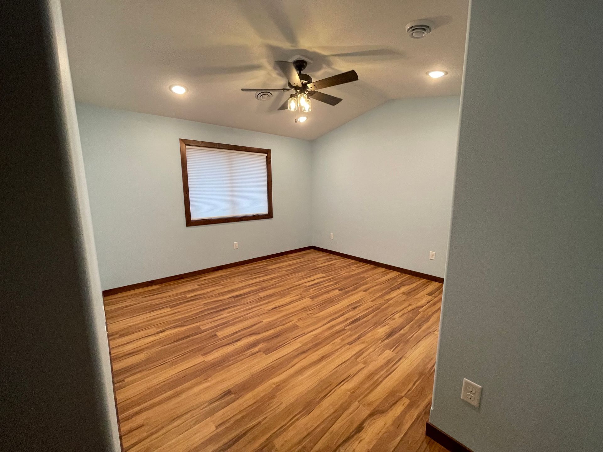Empty bedroom with wood floor, blue walls, a window with blinds, and a ceiling fan.