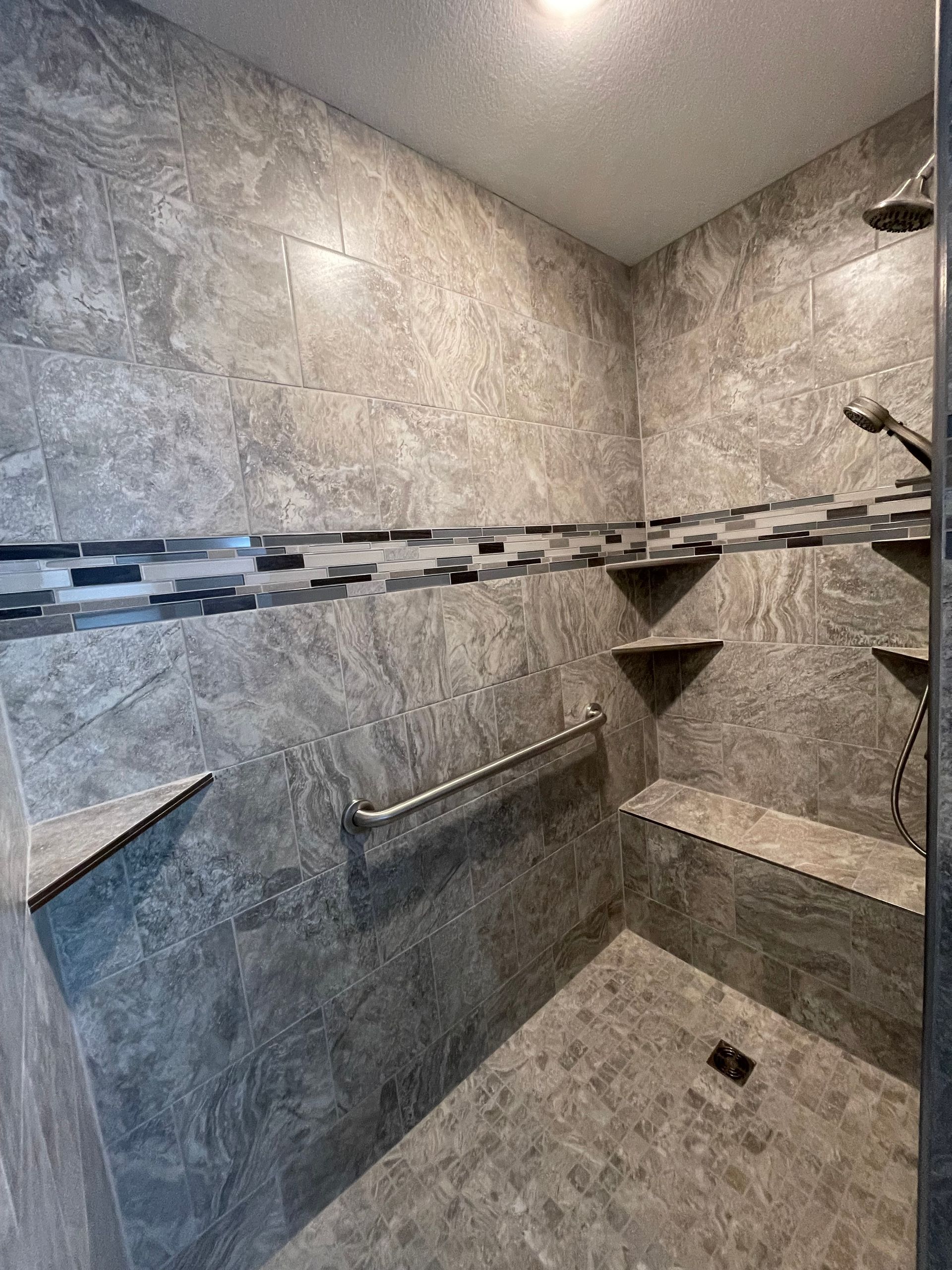 Gray tiled shower with shelves, a bench, and a grab bar.