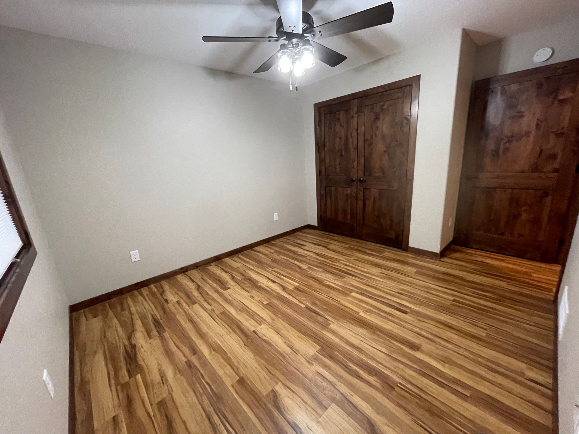 Empty bedroom with wood-look flooring, light walls, wooden closet doors, and a ceiling fan.