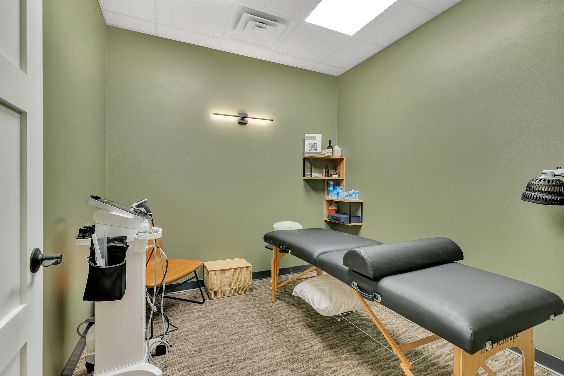 Therapy room with massage table, medical equipment, shelves. Olive walls, neutral floor.