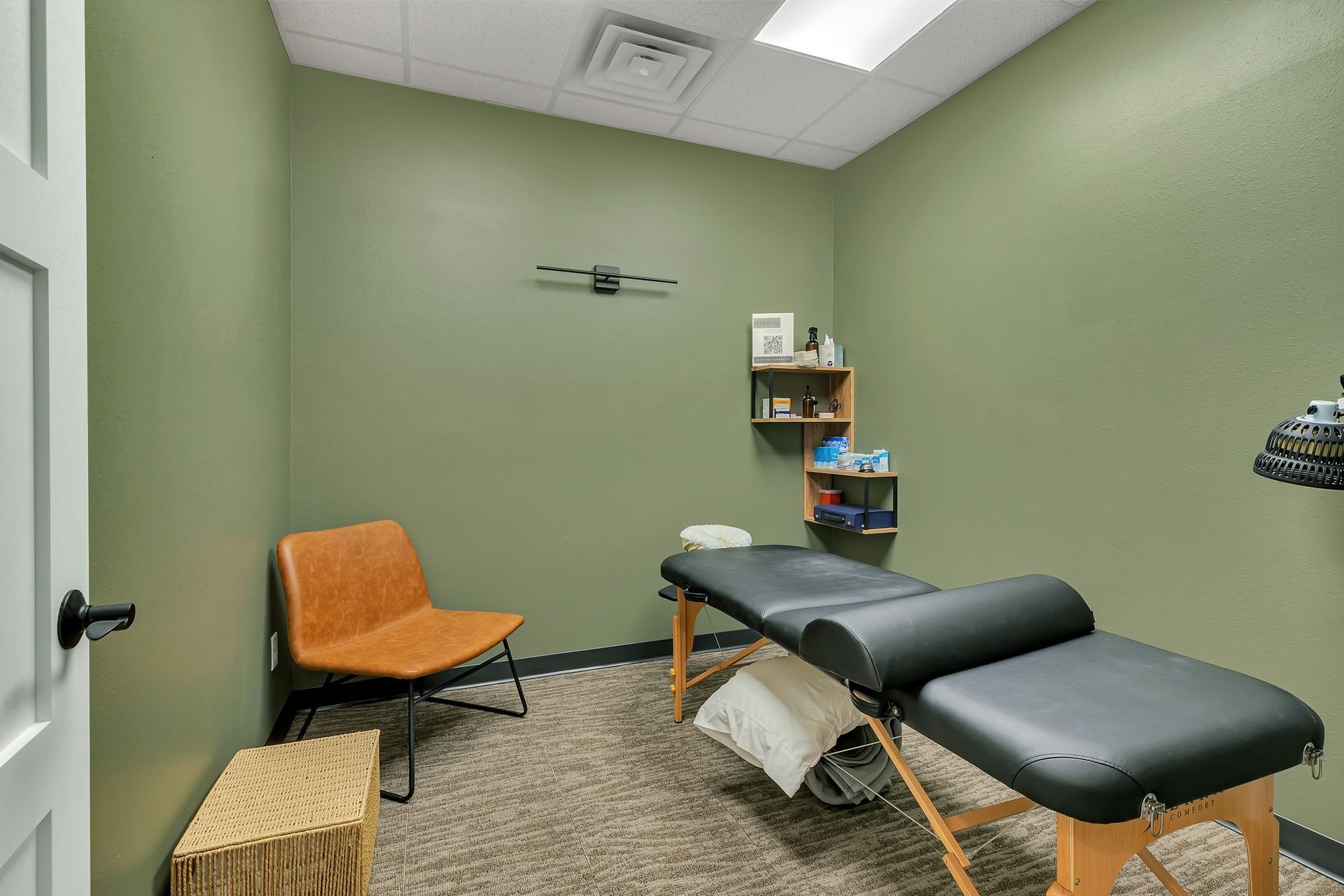 Small medical examination room with olive green walls, examination table, chair, and shelves.