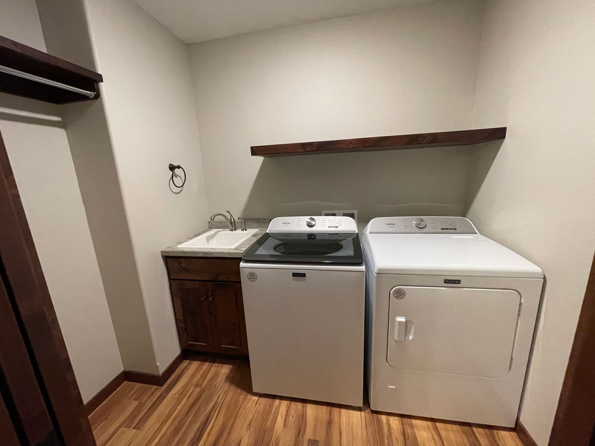 Laundry room with a washer, dryer, sink, and wooden shelf. Brown cabinets and flooring.