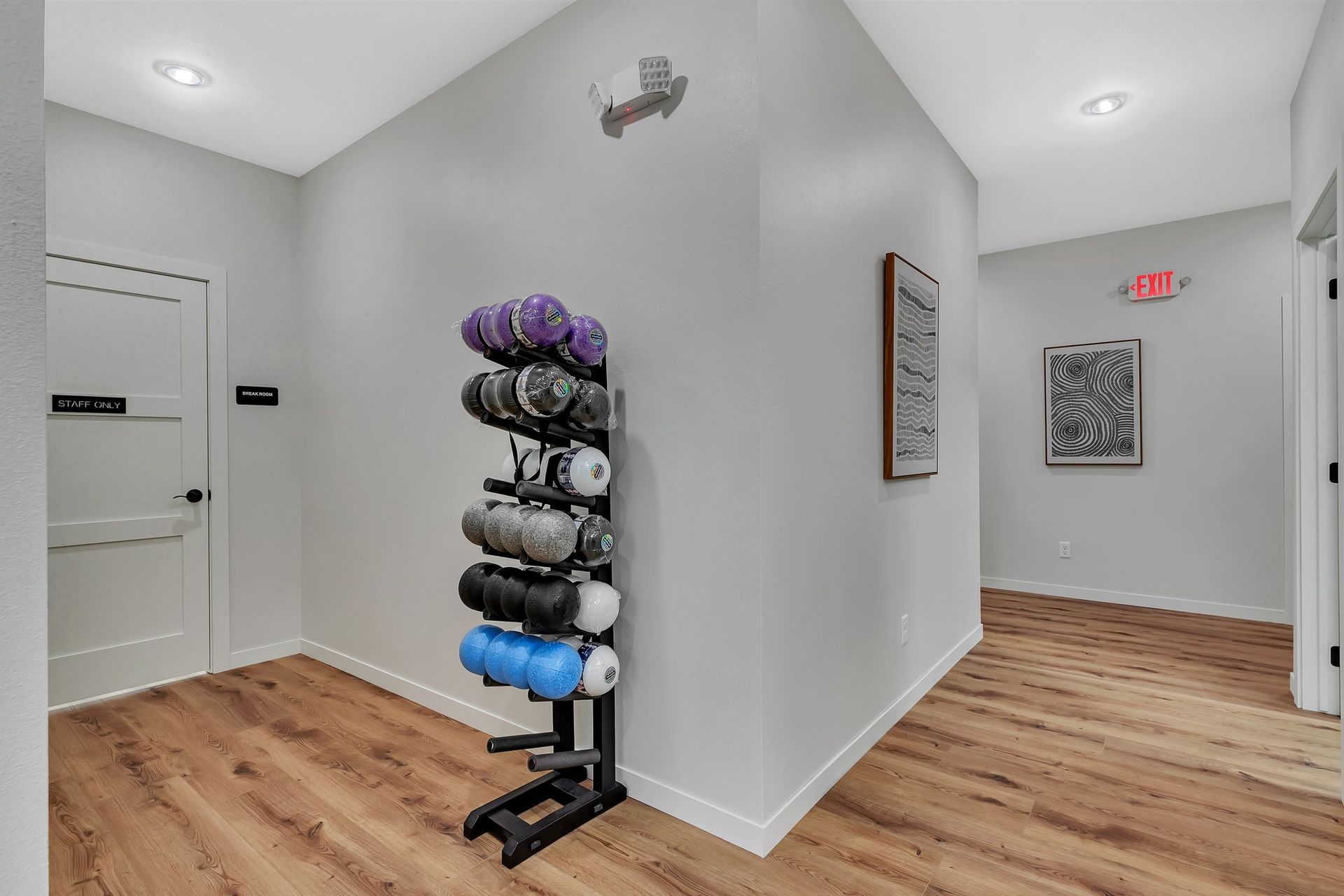 Hallway with wood flooring, weights on rack, framed art, and exit sign.