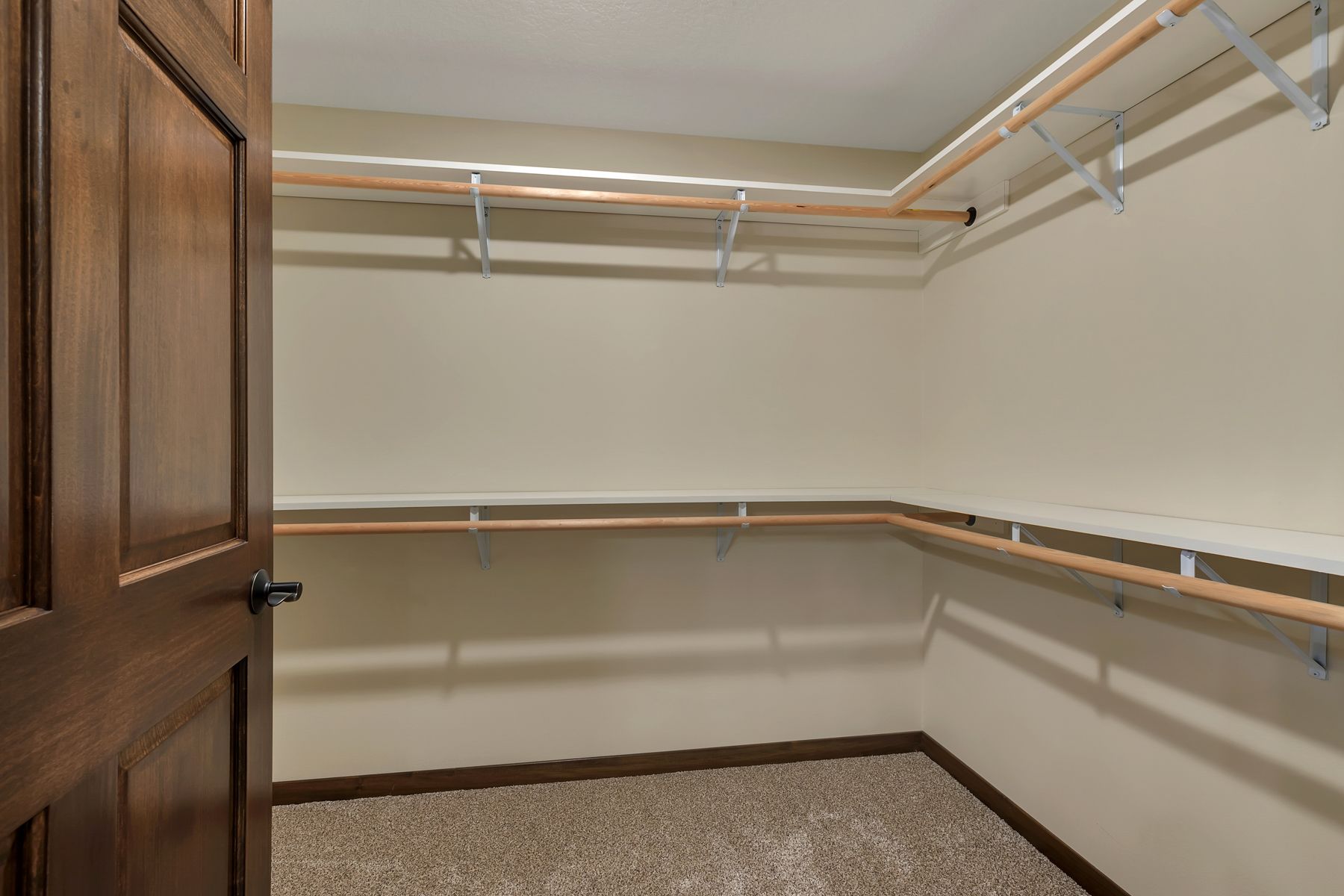 Empty walk-in closet with shelves and rods, beige walls, brown door, and carpet.