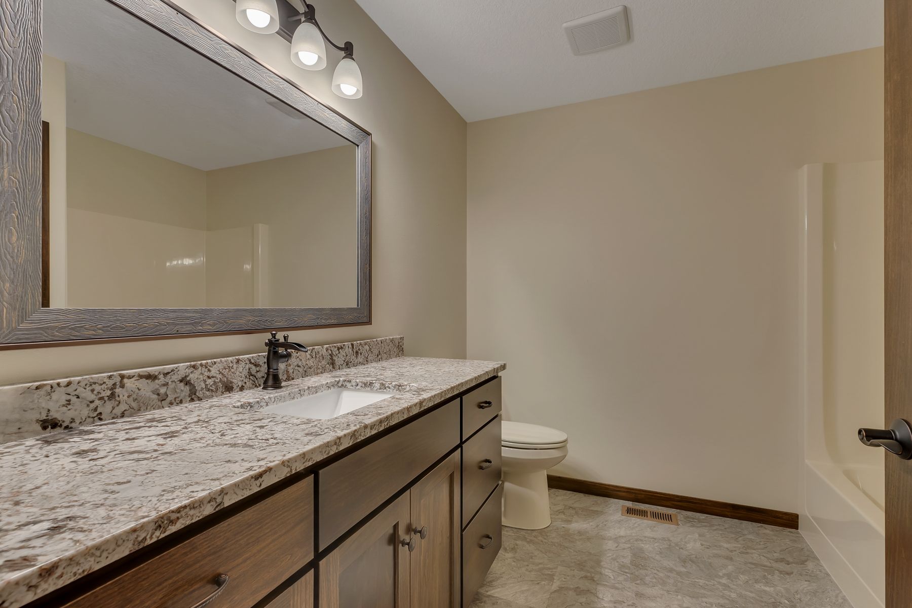 Bathroom with vanity, mirror, toilet, and shower. Tan walls, granite countertop, brown cabinets, and grey floor.