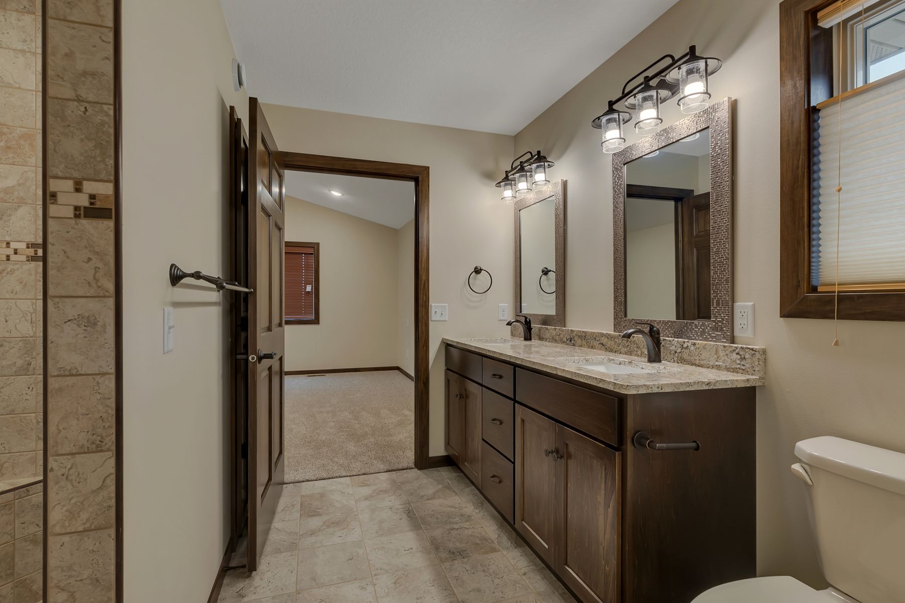 Bathroom with double vanity, two mirrors, and a toilet. Light brown tile floor and beige walls.