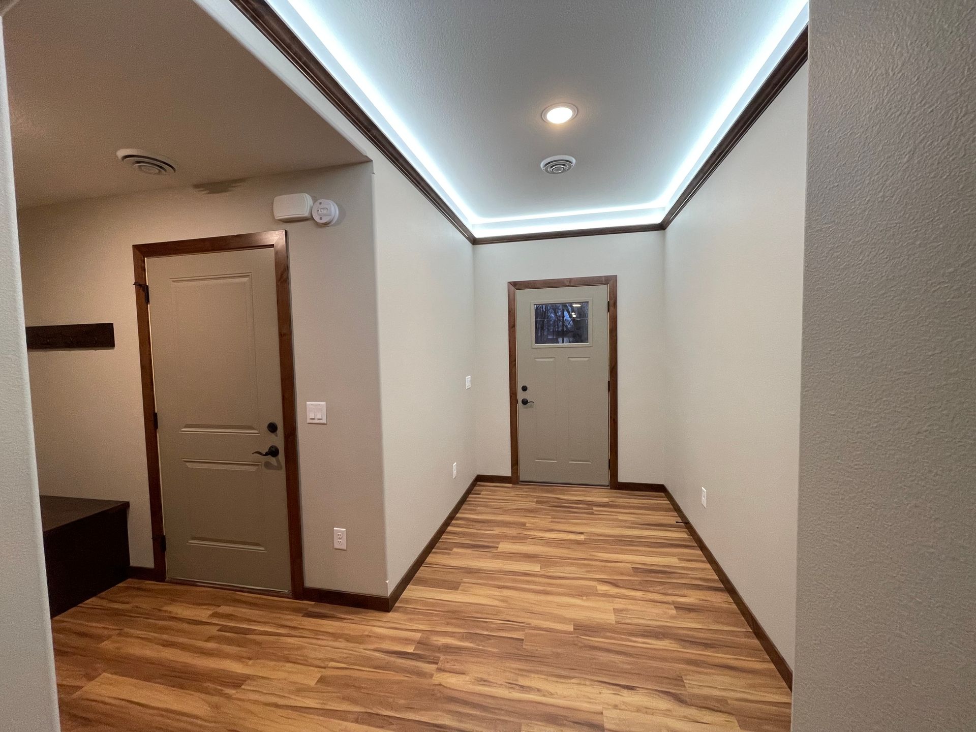 Hallway with wood-look flooring, two tan doors with brown trim, and a white ceiling with ambient lighting.