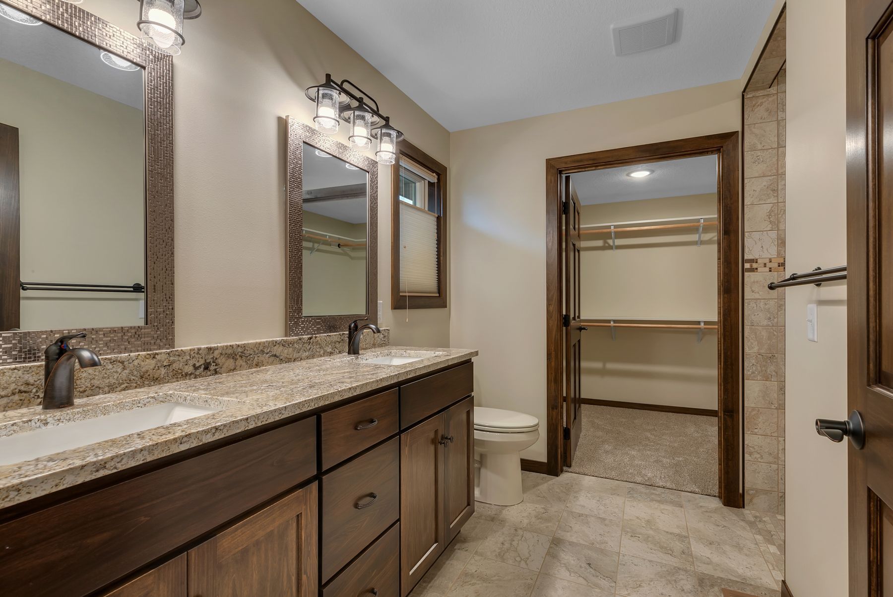 Bathroom with double vanity, framed mirrors, and a walk-in closet. Brown cabinets, beige walls, and light floor.
