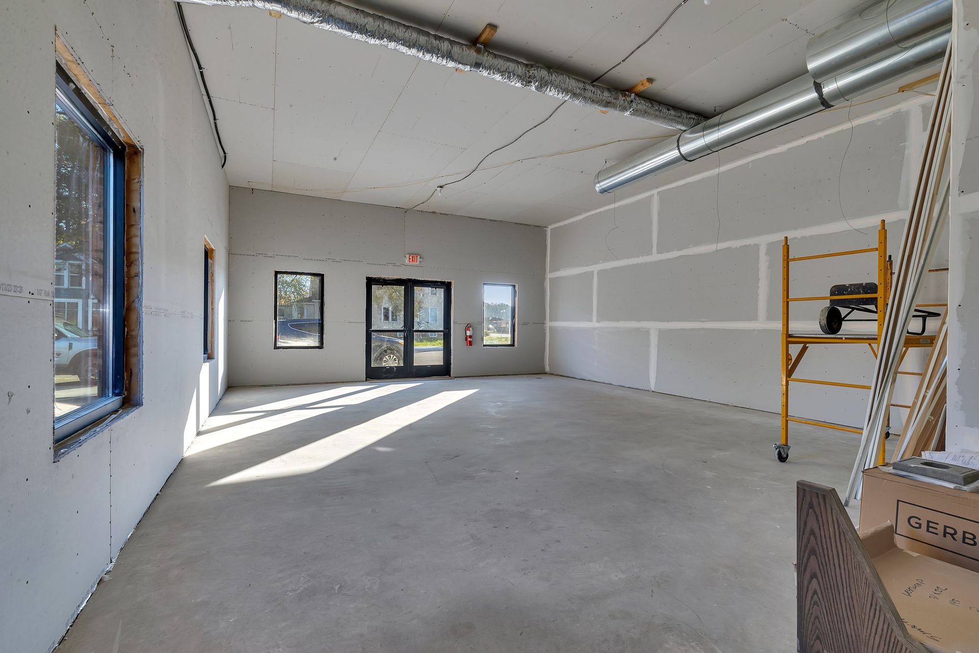 Empty room under construction with concrete floor, drywall, windows, and scaffolding.