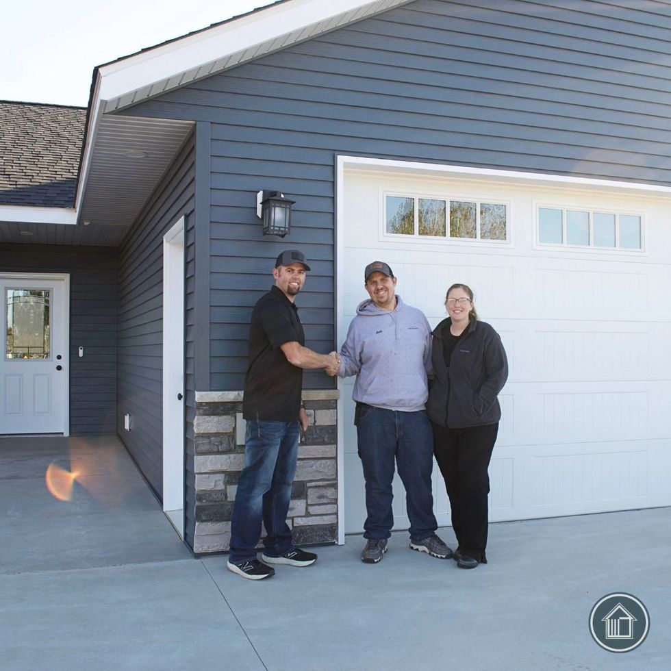 Three people standing outside a gray house beside a white garage door, one shaking hands with another.