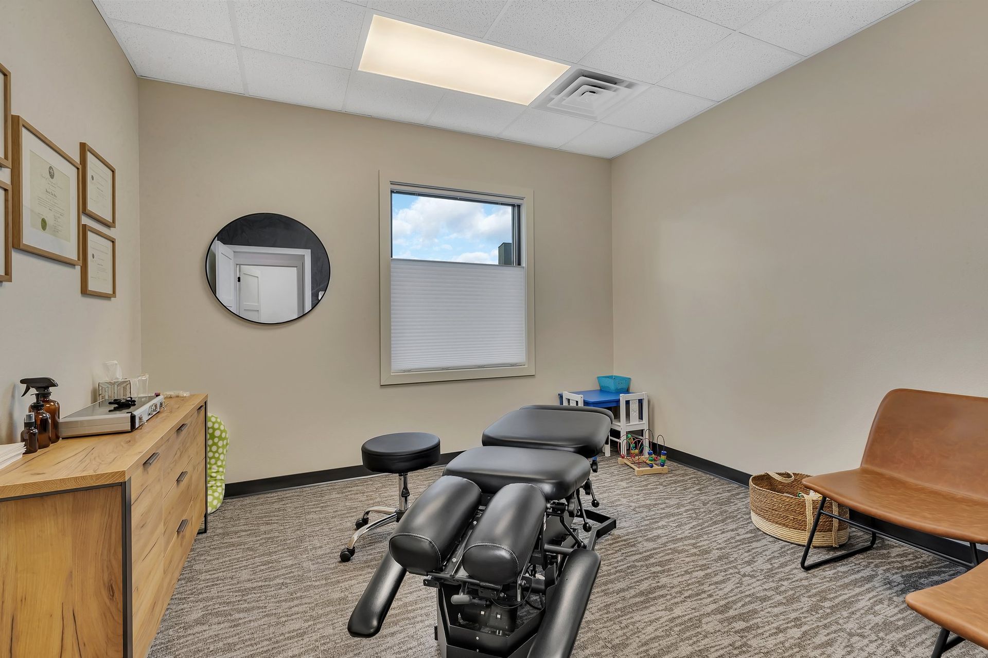 Chiropractor's office: beige walls, treatment table, waiting area with brown seating, small window, and round mirror.