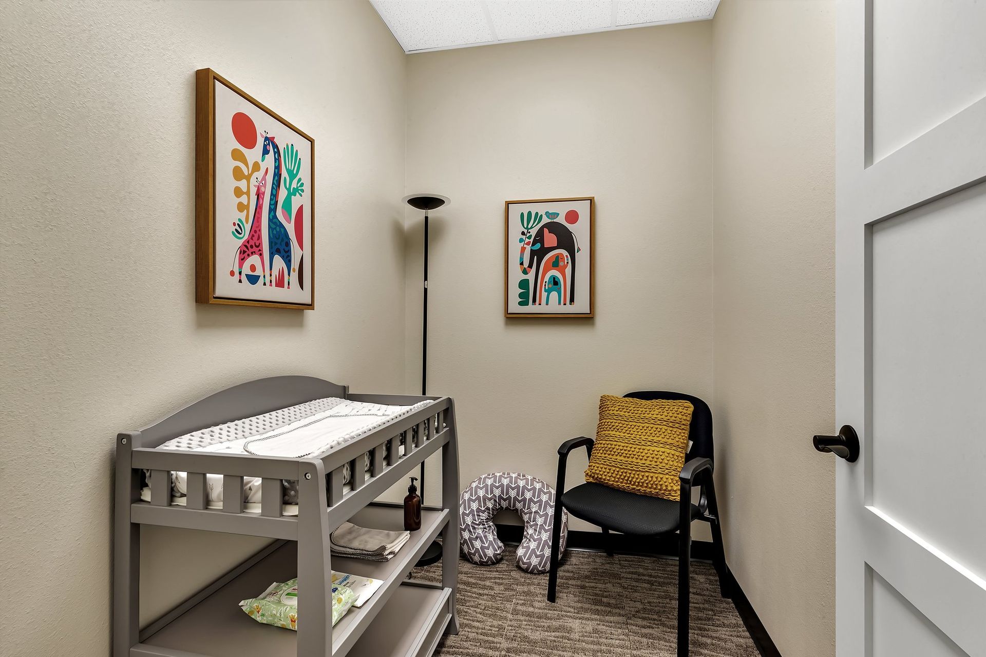 A baby changing room with gray changing table, black chair, and colorful art on the walls.