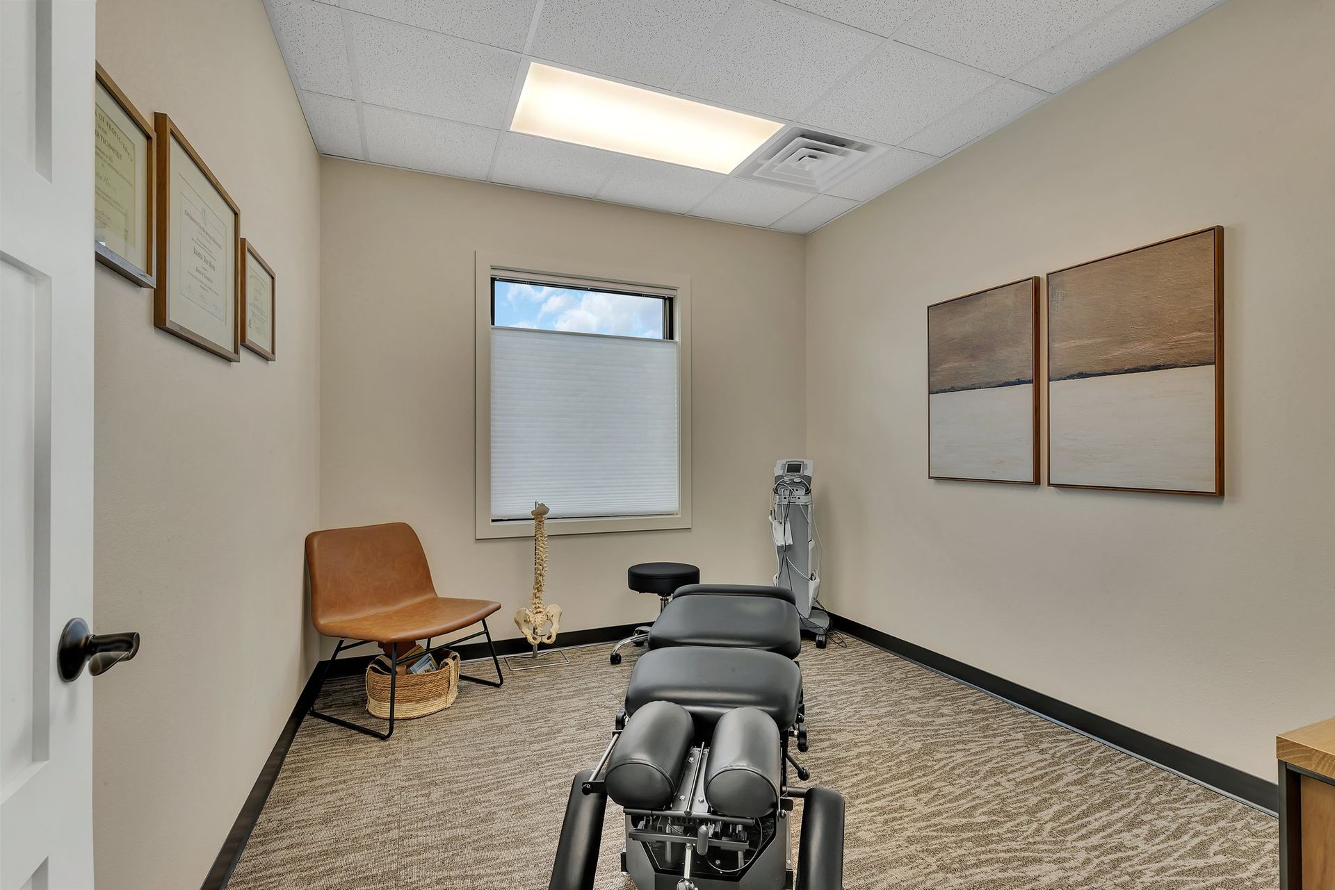 Chiropractor's office interior: beige walls, chiropractic table, small window, art, chair, skeleton model, and door.