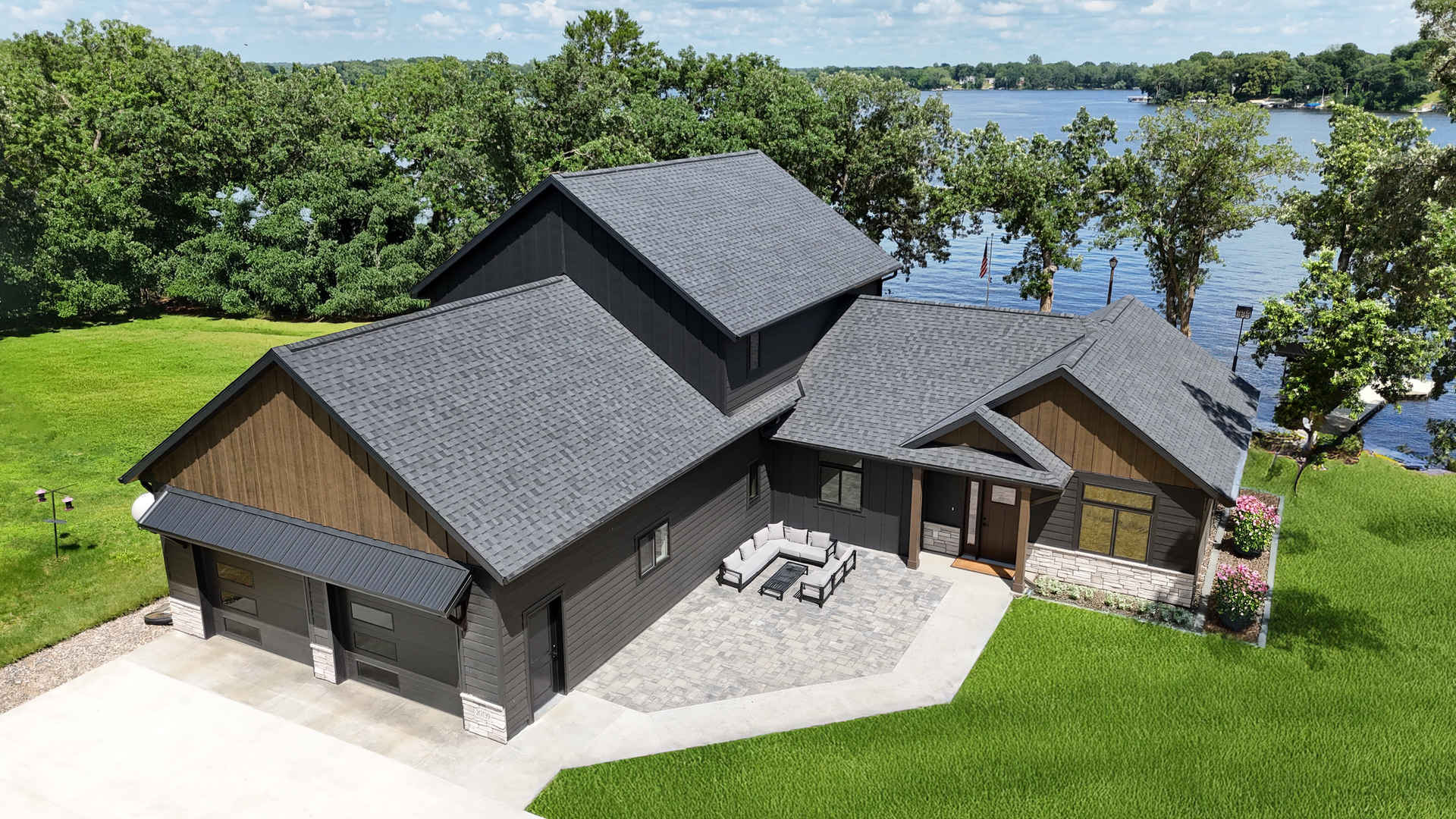 Aerial view of a modern dark-gray house with patio, surrounded by green lawn and trees near a lake