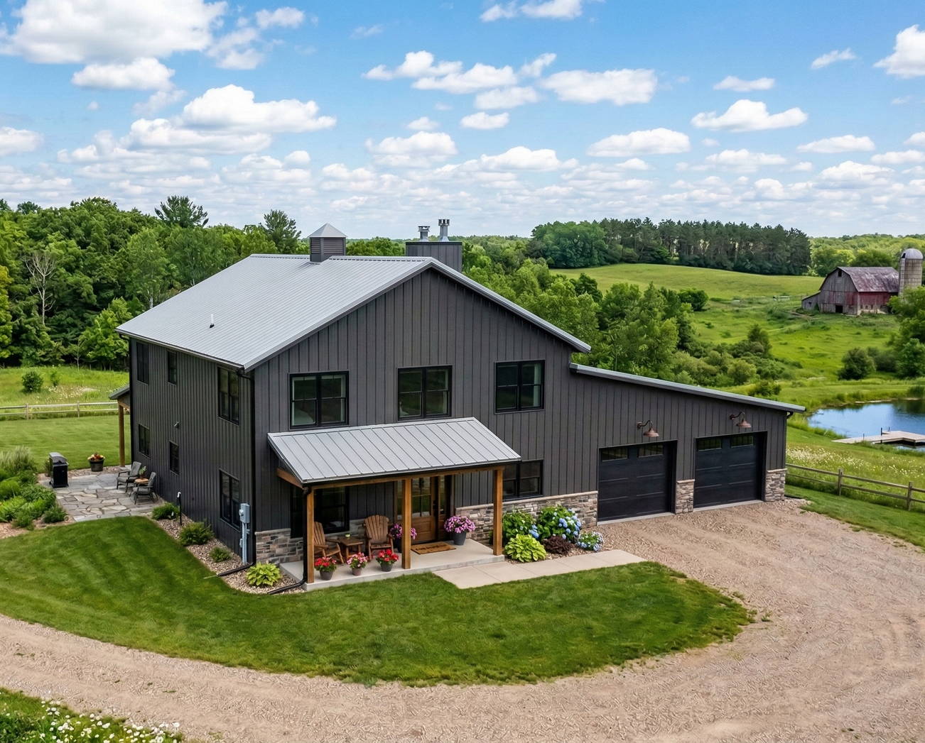 Dark gray farmhouse with a porch and gravel driveway beside a green field and pond under a blue sky