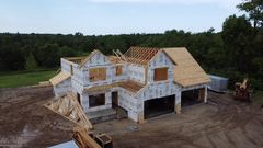 Partially built two-story house with exposed framing and roof sheathing on a dirt lot, surrounded by trees