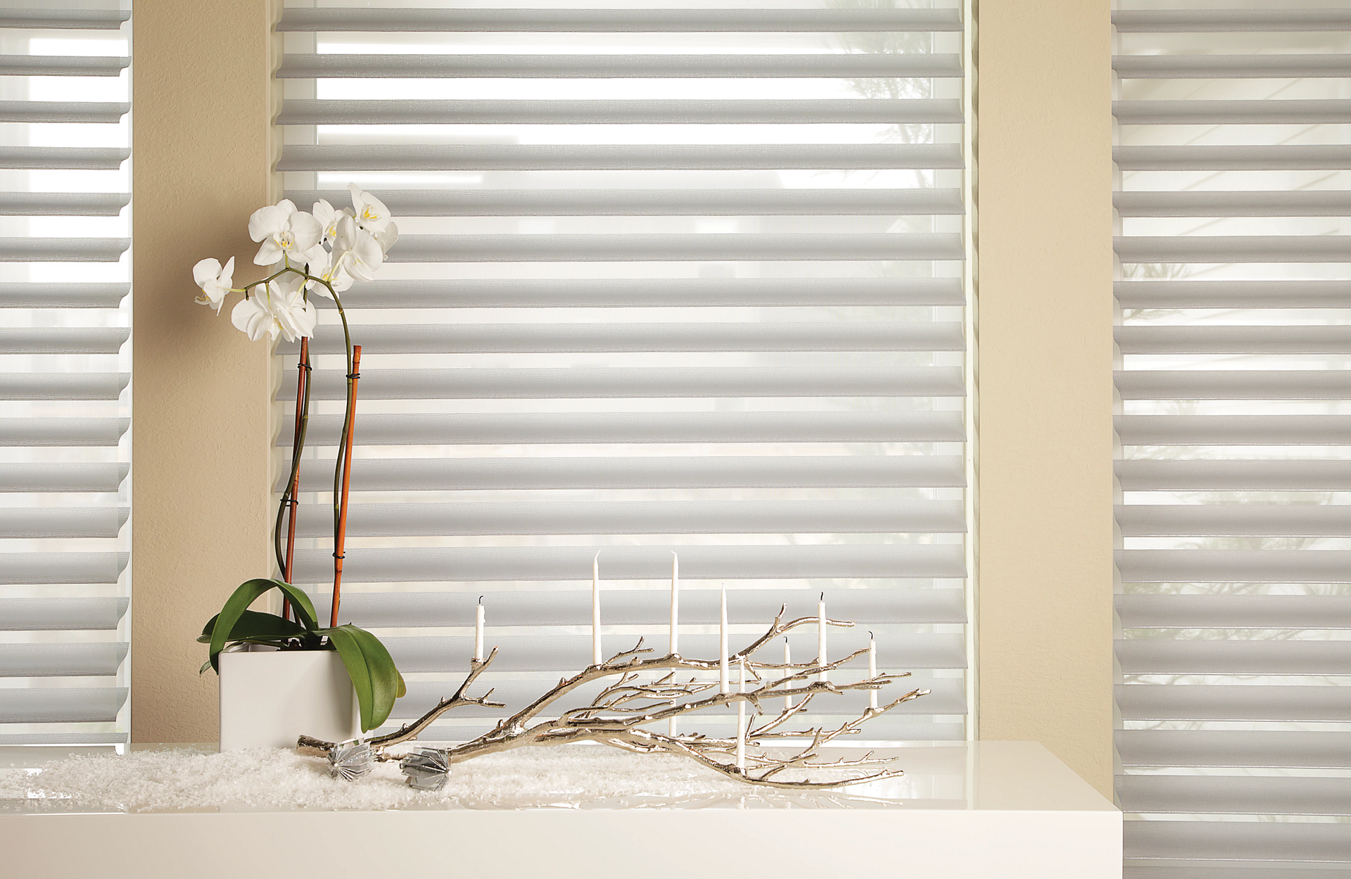 A vase of flowers and candles on a table in front of a window with blinds.