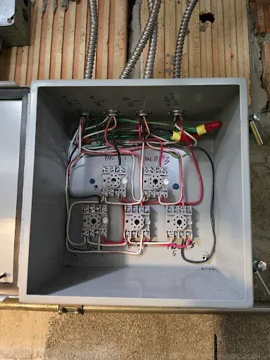 Open electrical box with wires connected to multiple relays against a wood and concrete wall.
