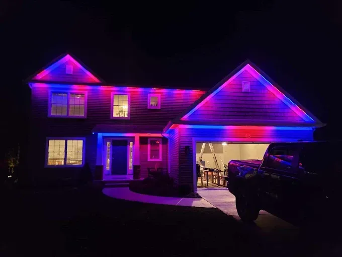 House at night, illuminated with blue and red lights along the rooflines and around the garage, a truck parked nearby.