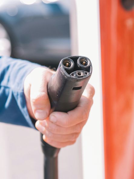 Hand holding an electric vehicle charging connector, red and white charging station in background.