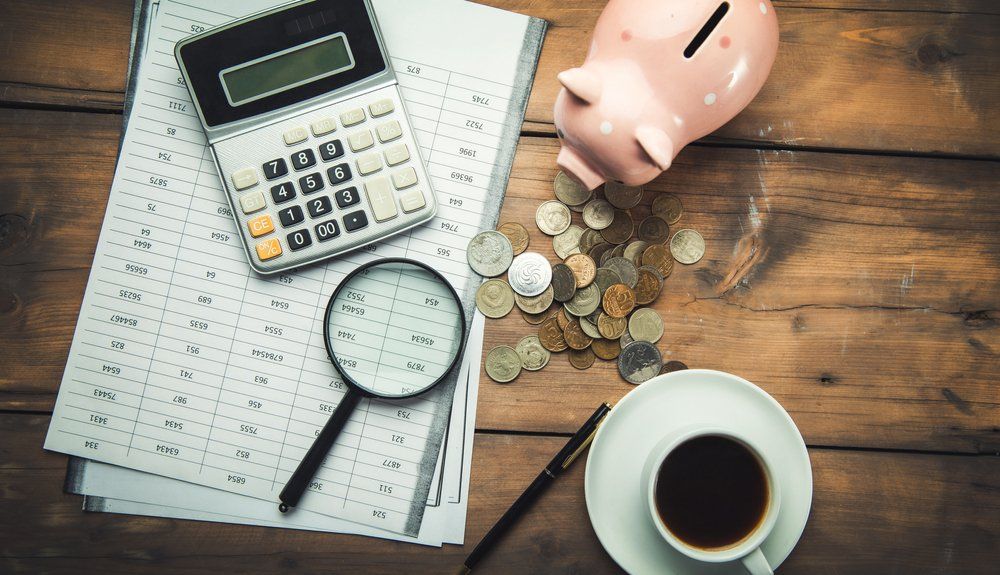 Calculator, magnifying glass, paperwork, coins, piggy bank, coffee cup, and pen on a wooden table.