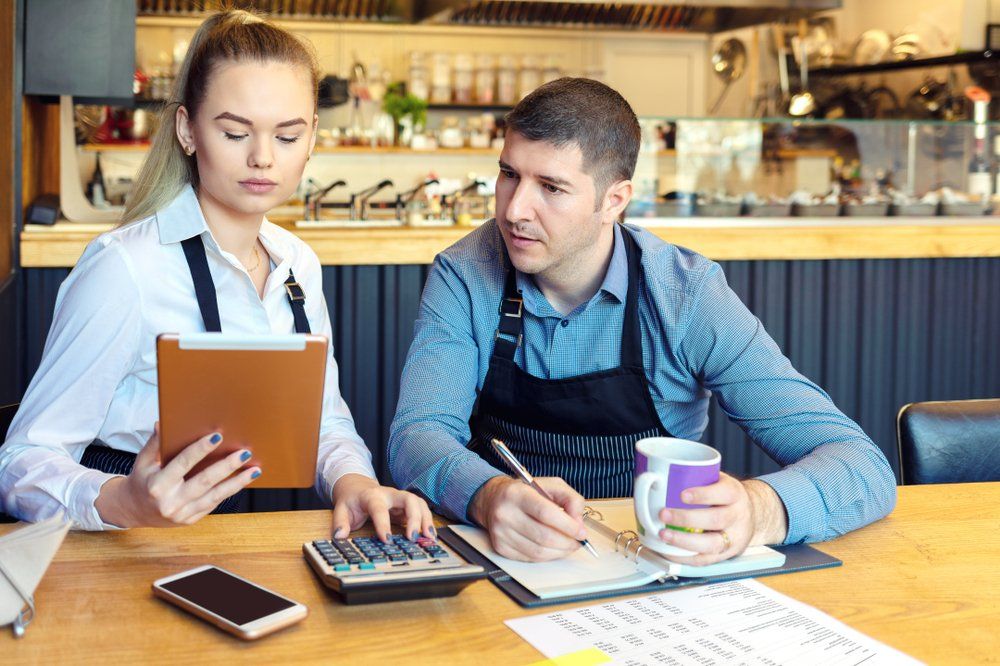 Woman and man reviewing finances at a restaurant table. Woman holds tablet, man writes in a notebook with a calculator and mug.